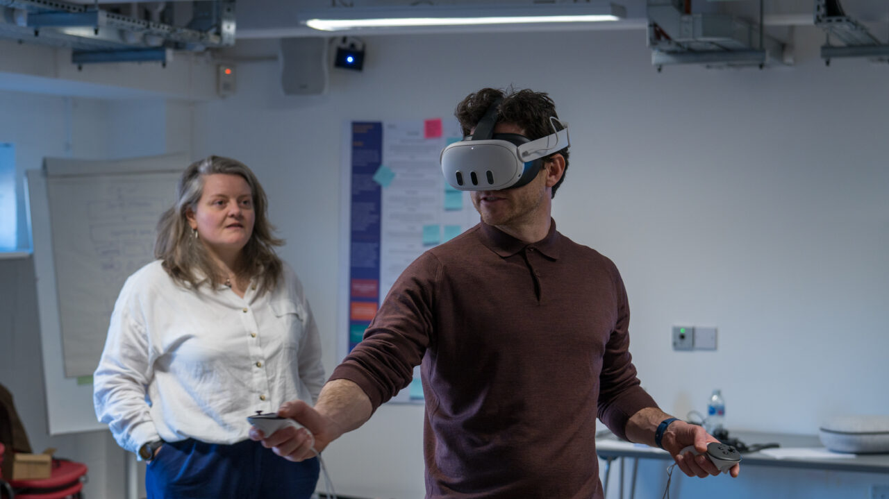 A man wearing a virtual reality headset and holding controllers is using VR in a room, while a woman stands nearby watching him. Mae dyn sy'n gwisgo clustffon realiti rhithwir ac yn dal rheolyddion yn defnyddio realiti rhithwir mewn ystafell, tra bod menyw yn sefyll gerllaw yn ei wylio.