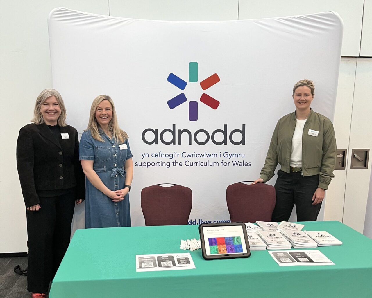 Three women stand smiling behind an exhibition table with a green tablecloth and branding for "adnodd", an organisation supporting the Curriculum for Wales. The backdrop and materials on the table promote educational resources. Mae tair menyw yn sefyll yn gwenu y tu ôl i fwrdd arddangos gyda lliain bwrdd gwyrdd a brandio ar gyfer "adnodd", sefydliad sy'n cefnogi Cwricwlwm Cymru. Mae'r cefndir a'r deunyddiau ar y bwrdd yn hyrwyddo adnoddau addysgol.