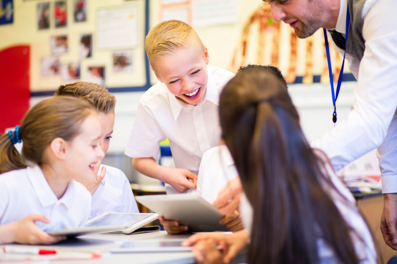 A group of primary school children in uniform smiling and using tablets in a classroom, with a teacher engaging with them. Grŵp o blant ysgol gynradd mewn gwisg ysgol yn gwenu ac yn defnyddio tabledi mewn ystafell ddosbarth, gydag athro yn ymgysylltu â nhw.