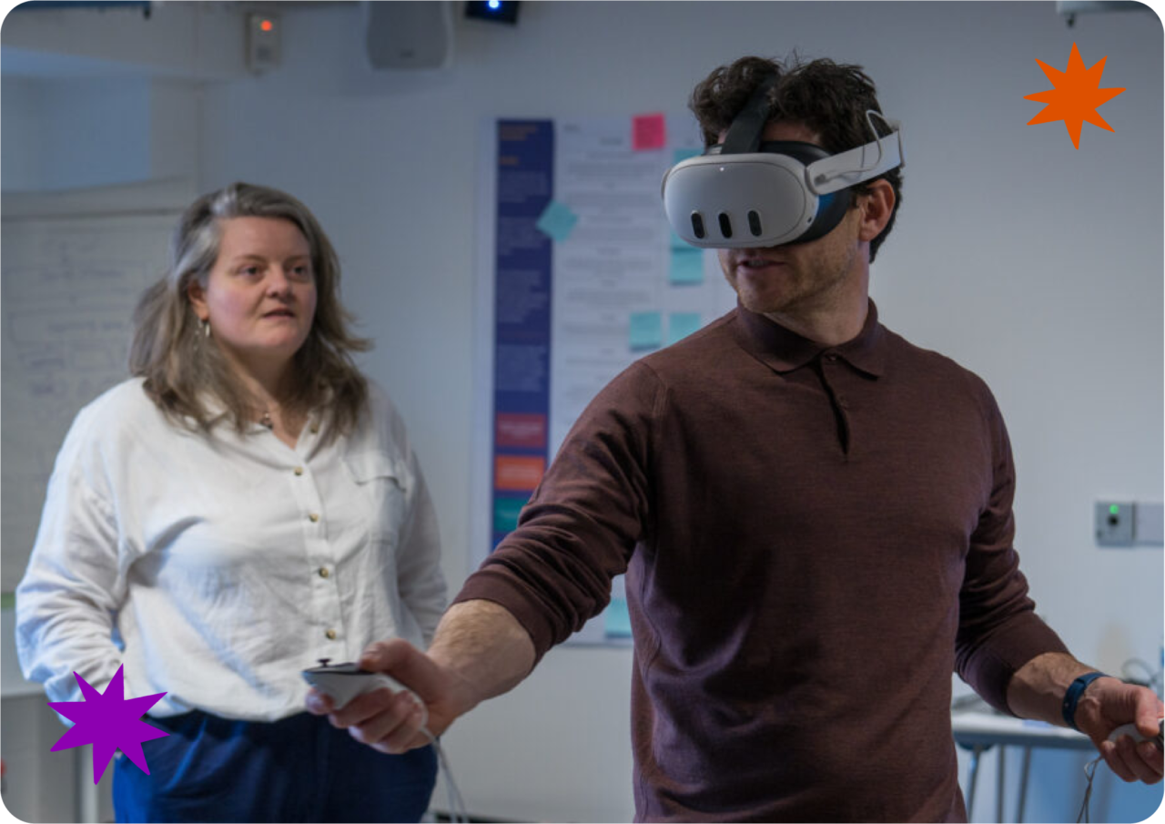 A man wearing a virtual reality headset and holding controllers is using VR in a room, while a woman stands nearby watching him. Mae dyn sy'n gwisgo clustffon realiti rhithwir ac yn dal rheolyddion yn defnyddio realiti rhithwir mewn ystafell, tra bod menyw yn sefyll gerllaw yn ei wylio.