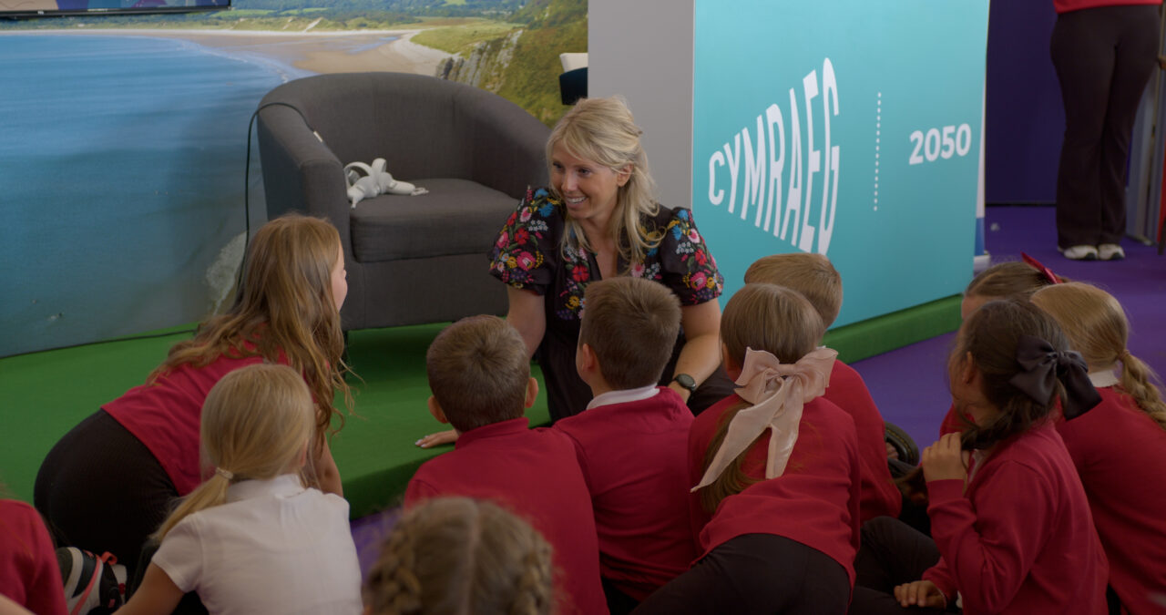 A group of primary school children in uniform smiling and using tablets in a classroom, with a teacher engaging with them. Grŵp o blant ysgol gynradd mewn gwisg ysgol yn gwenu ac yn defnyddio tabledi mewn ystafell ddosbarth, gydag athro yn ymgysylltu â nhw.