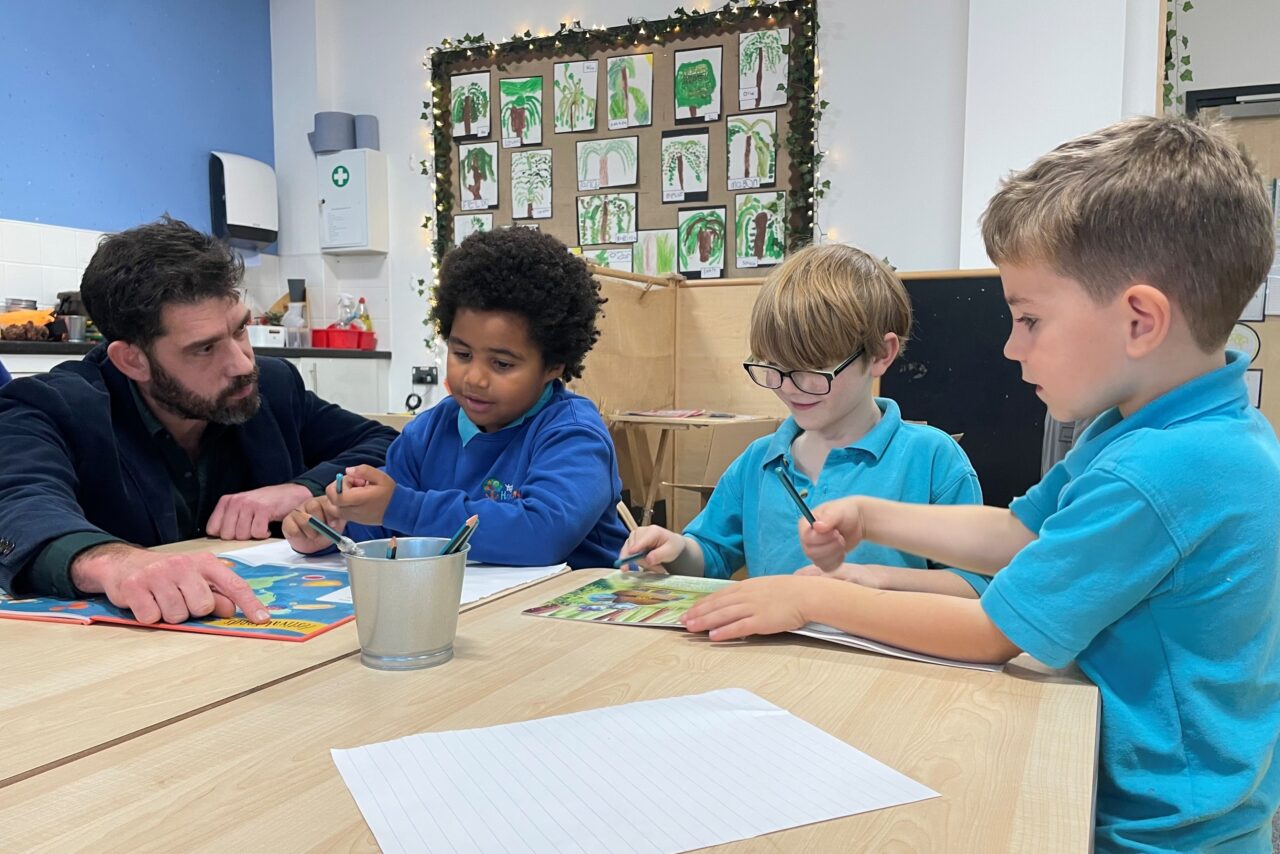 An adult sits with three young pupils at a classroom table, looking at books and chatting as they draw.