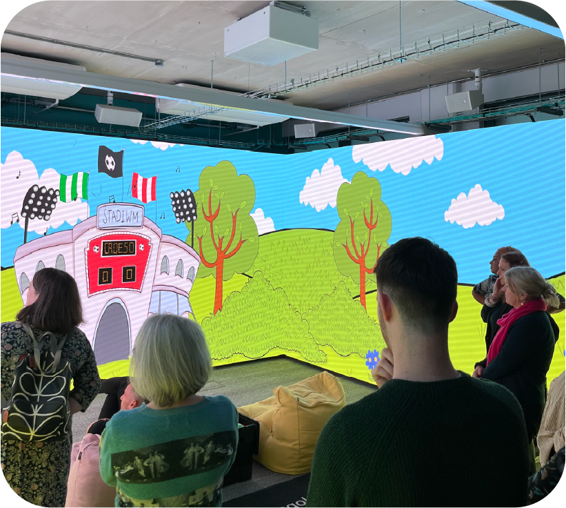 A group of people stand and sit facing a large curved digital screen showing a colourful cartoon stadium, trees, and a scoreboard. | Mae grŵp o bobl yn sefyll ac yn eistedd yn wynebu sgrin ddigidol fawr grom sy’n dangos stadiwm cartŵn lliwgar, coed, a sgorfwrdd.