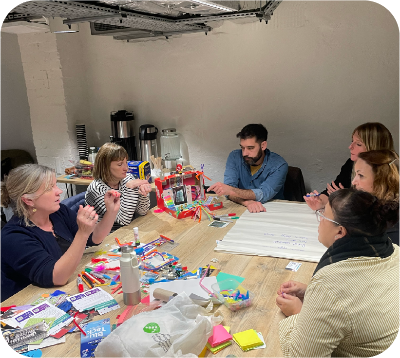 A group of adults sit around a table in a workshop setting, using craft materials and markers while discussing ideas and writing on a large sheet of paper | Mae grŵp o oedolion yn eistedd o amgylch bwrdd mewn lleoliad gweithdy, yn defnyddio deunyddiau crefft a beiros tra’n trafod syniadau ac yn ysgrifennu ar ddalen fawr o bapur.
