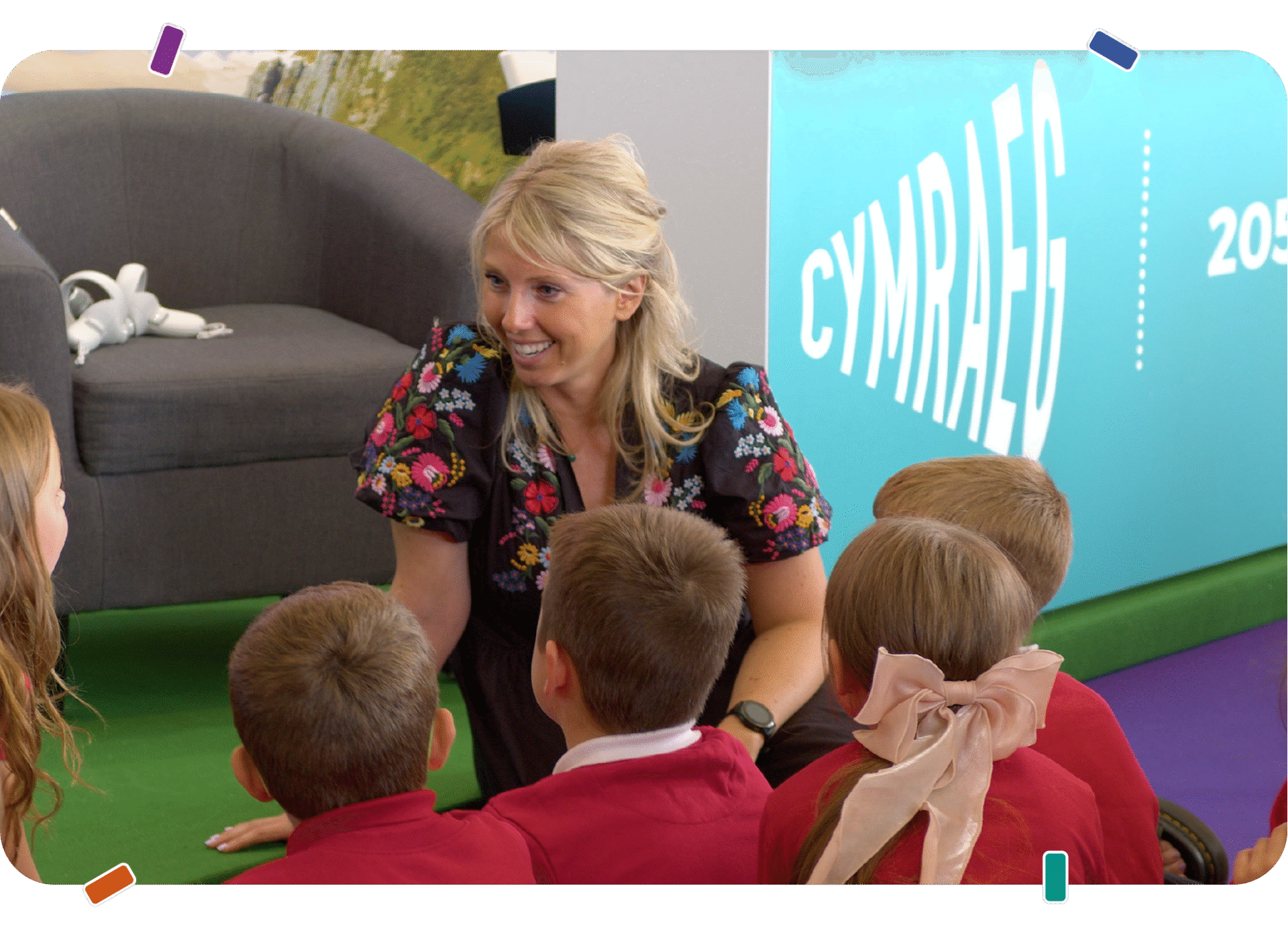 A woman kneels and talks with a small group of young schoolchildren sitting on the floor in front of a display with the word “Cymraeg”. | Mae menyw yn penlinio ac yn siarad â grŵp bach o blant ysgol ifanc sy’n eistedd ar y llawr o flaen arddangosfa gyda’r gair “Cymraeg”.