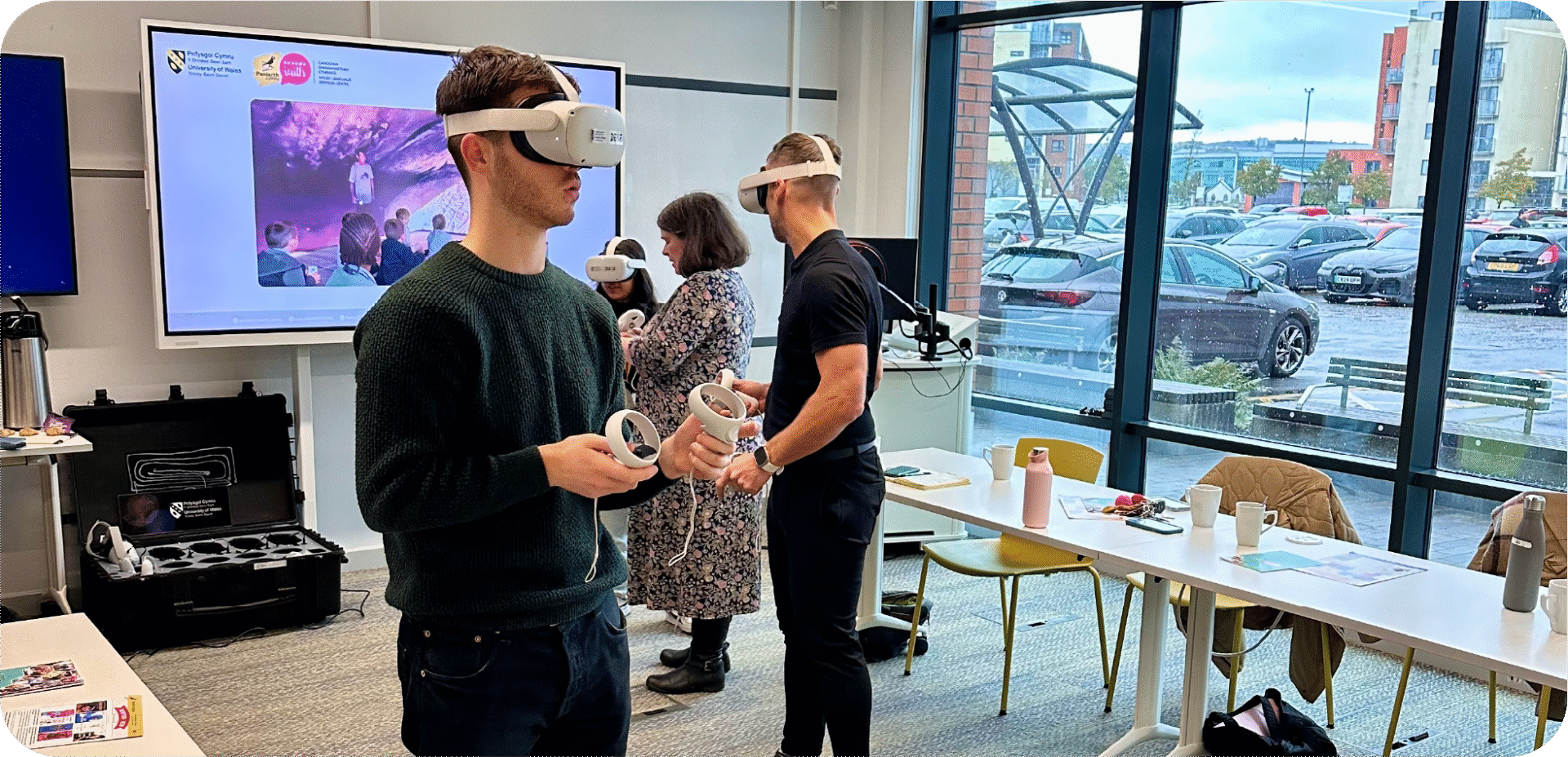 Three adults stand in a bright room using VR headsets and controllers, with tables, cups, and a large screen behind them. | Mae tri oedolyn yn sefyll mewn ystafell ddisglair yn defnyddio setiau realiti rhithwir a rheolwyr, gyda byrddau, cwpanau a sgrin fawr y tu ôl iddynt.