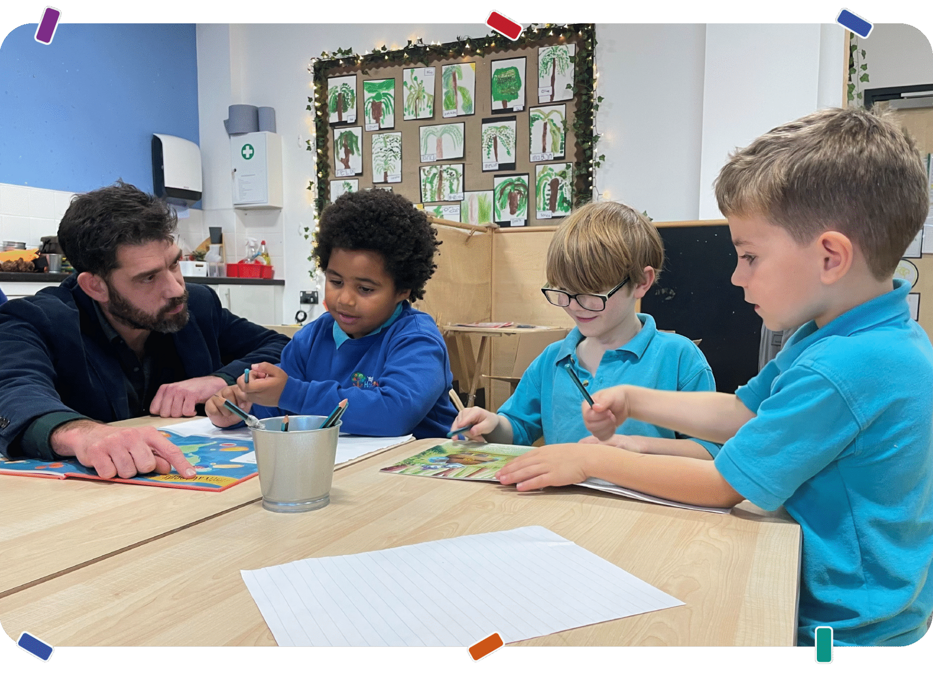 An adult sits at a table with four young children in a classroom, reading a book and drawing together with pencils and paper | Mae oedolyn yn eistedd wrth fwrdd gyda phedwar o blant ifanc mewn ystafell ddosbarth, yn darllen llyfr ac yn tynnu lluniau gyda phensiliau a phapur.