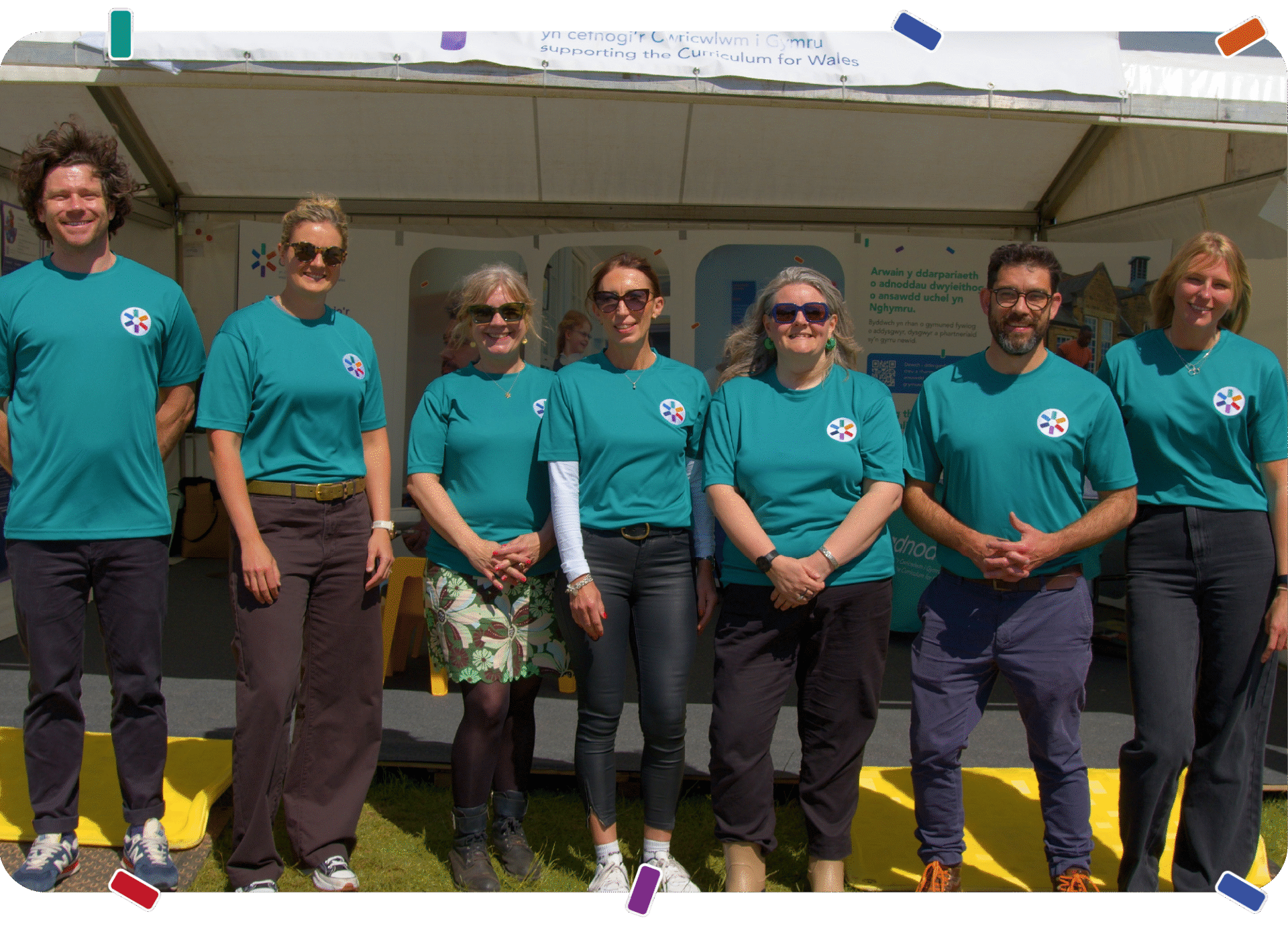 Seven members of the Adnodd team stand side by side outdoors in front of a stall, wearing matching Adnodd-branded T-shirts and smiling at the camera. | Mae saith aelod o dîm Adnodd yn sefyll ochr yn ochr y tu allan o flaen stondin, yn gwisgo crysau-T gyda logo Adnodd ac yn gwenu at y camera.